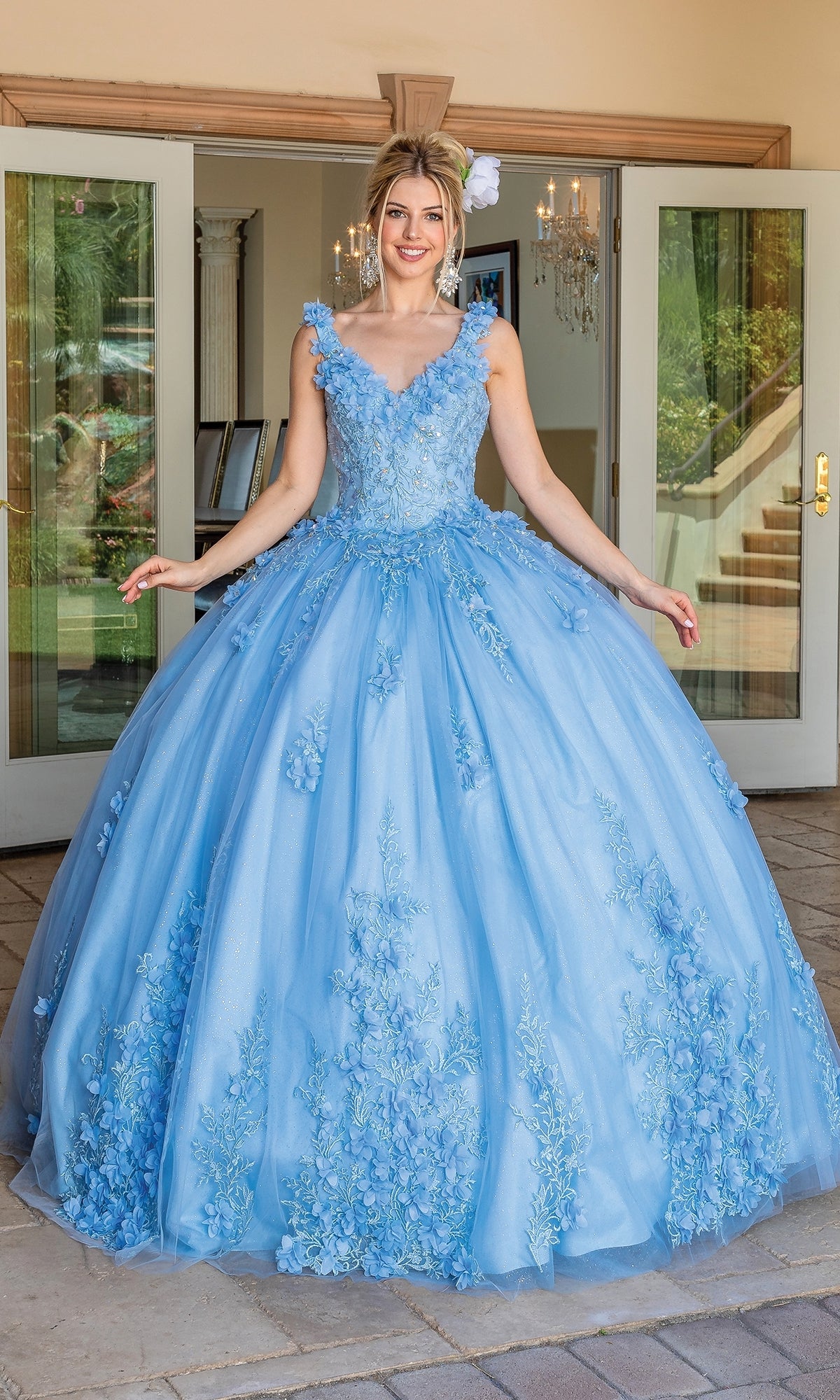 A woman stands outdoors, smiling in front of open glass doors, wearing the Quinceanera Dress 1623 by Dancing Queen—a light blue ball gown with floral appliqués and a voluminous skirt.